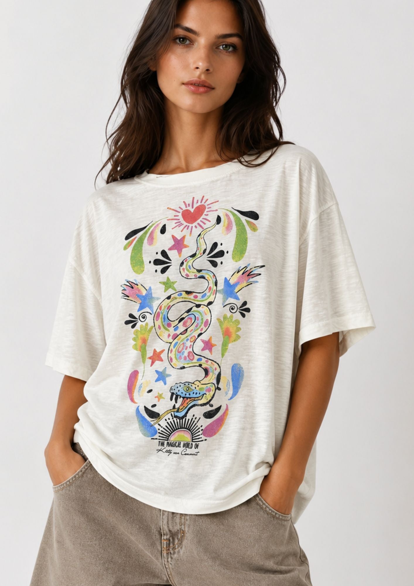 A woman wearing a cream-colored oversized t-shirt with a colorful snake graphic print, paired with light brown jeans, against a plain light gray background.