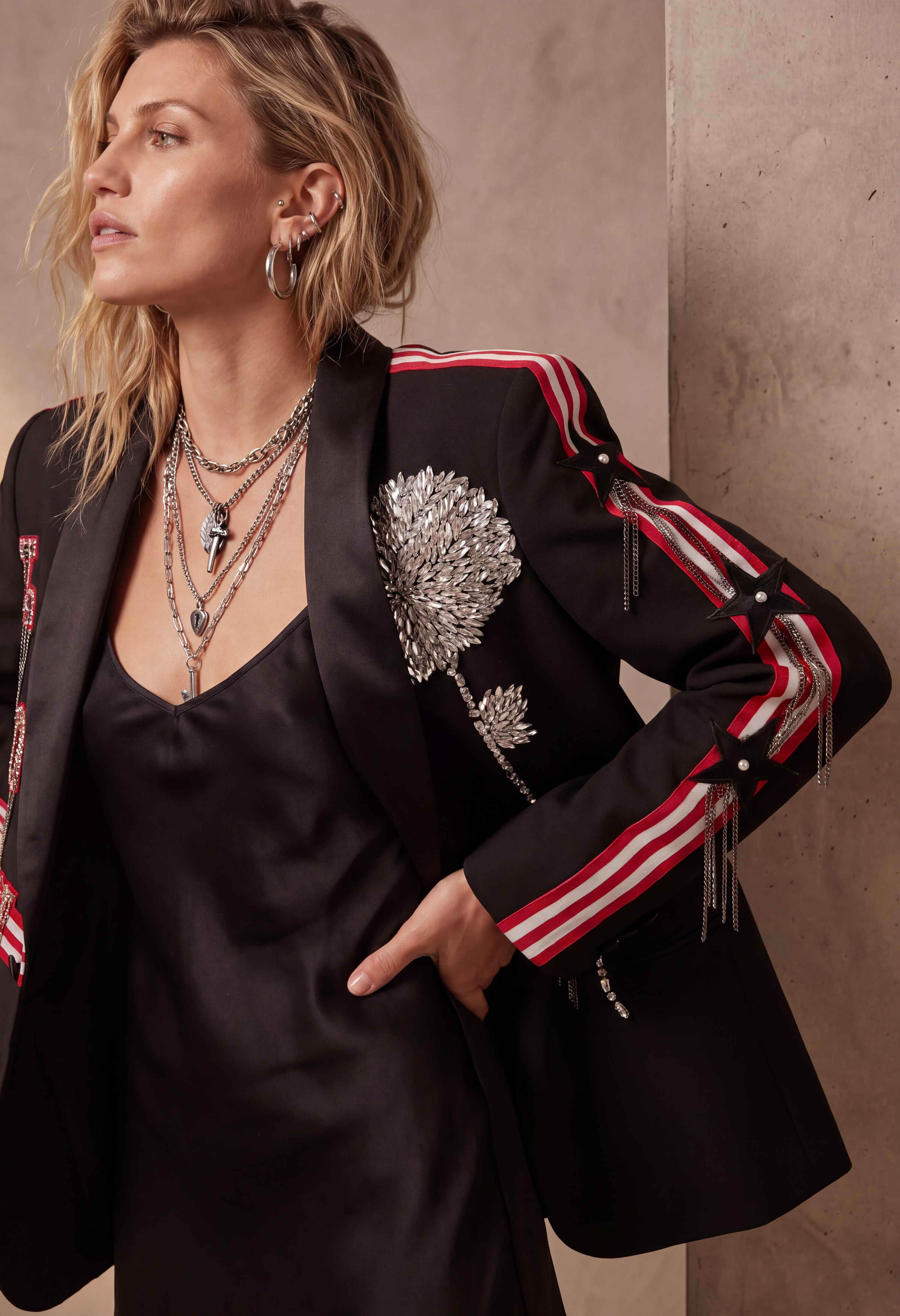 A woman wearing a black blazer with silver floral embroidery and red-and-white striped sleeve details, paired with a black satin slip dress and layered silver necklaces, posed against a textured beige wall.
