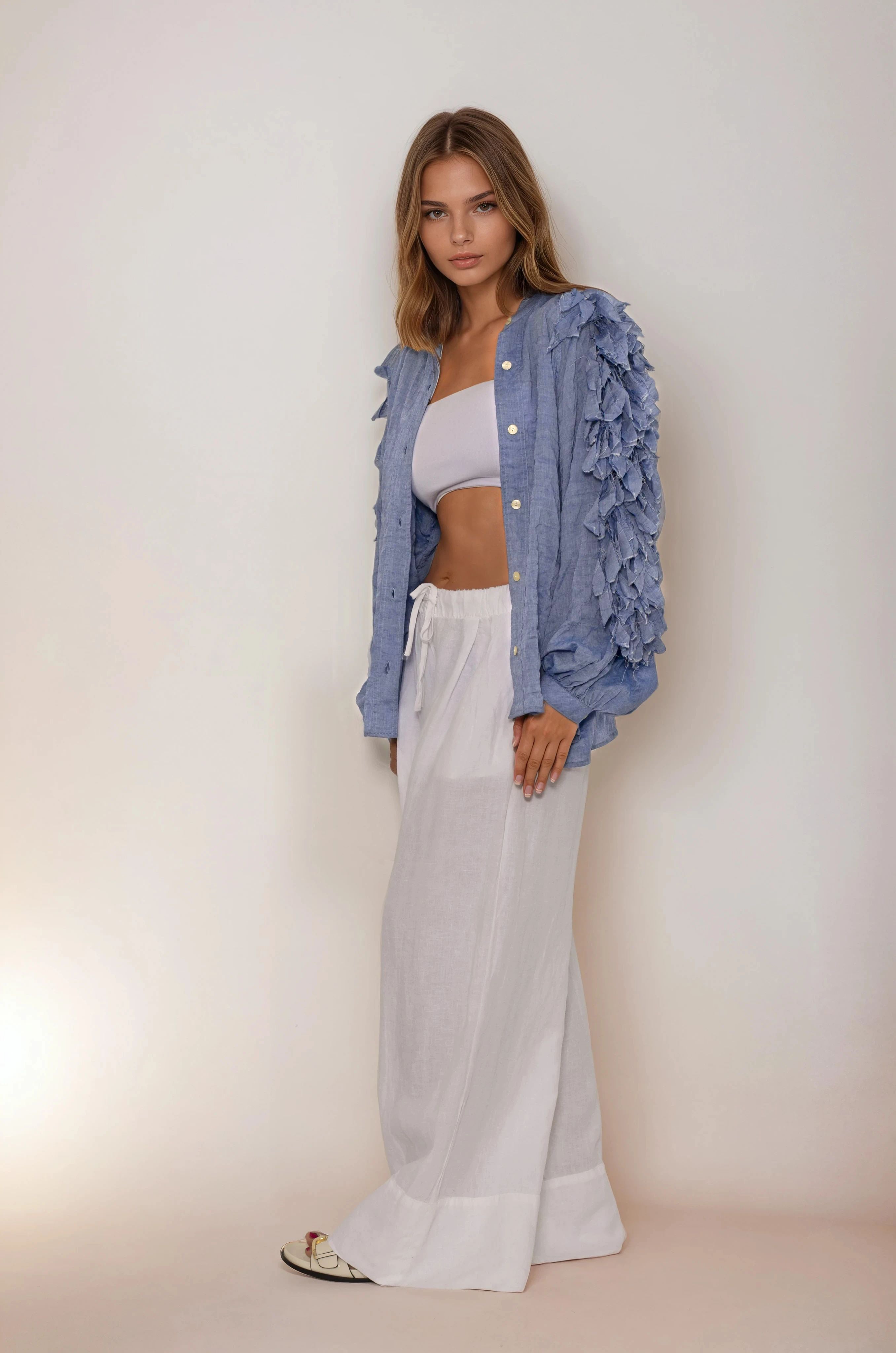 Woman wearing a blue ruffled jacket, white top, and wide-leg pants against a plain background