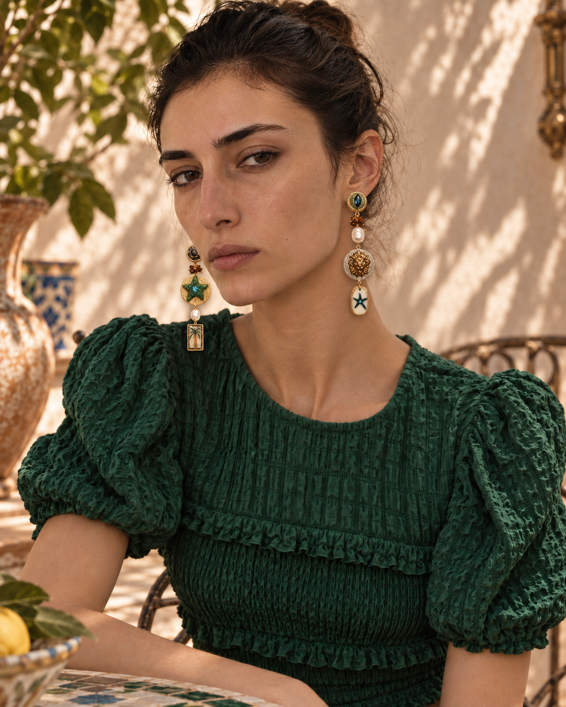 A woman wearing a dark green textured blouse and matching green statement earrings with gold, green, and white details, posed in a sunlit outdoor setting.