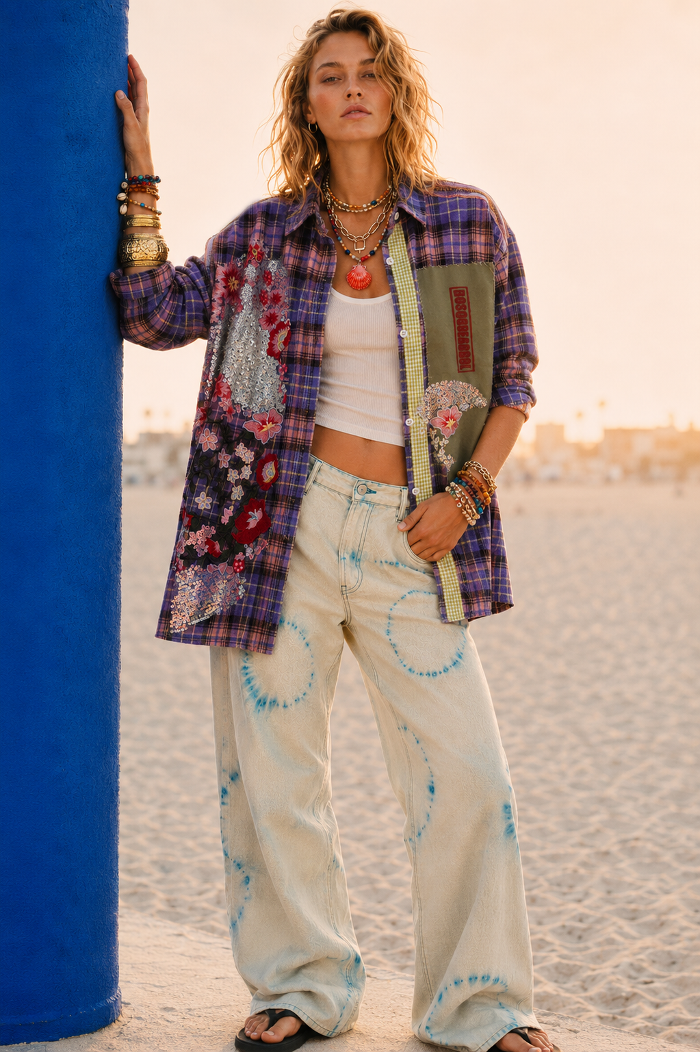 Woman wearing a colorful patchwork shirt and patterned pants standing against a blue pole.