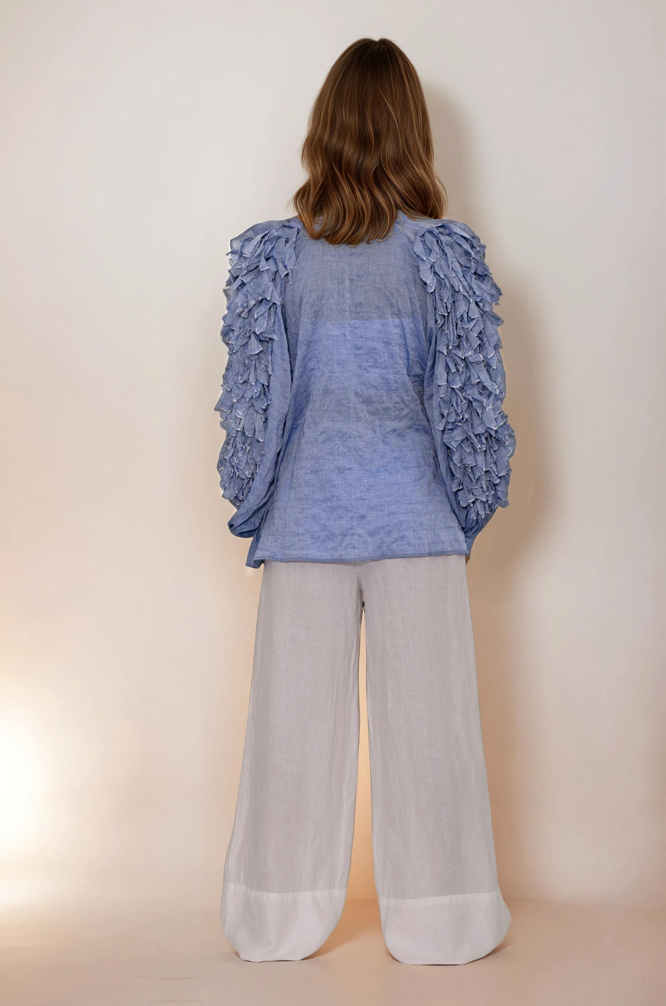 Person wearing a blue ruffled Claramonte top and light gray pants against a plain background