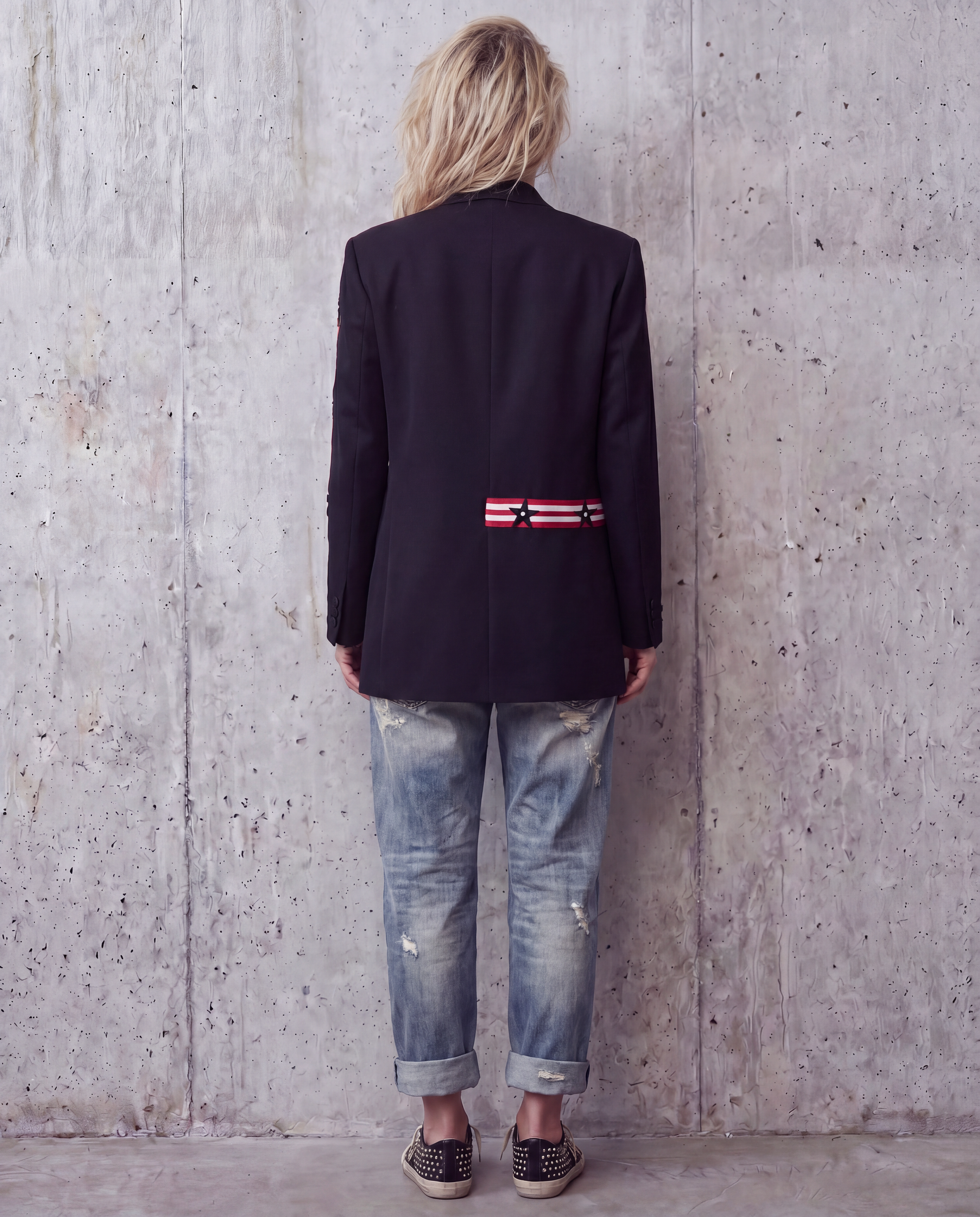 A woman viewed from behind wearing a dark navy blazer with a red, white, and black striped ribbon detail on the back, paired with distressed light-wash blue jeans and black-and-white sneakers, standing against a textured concrete wall.