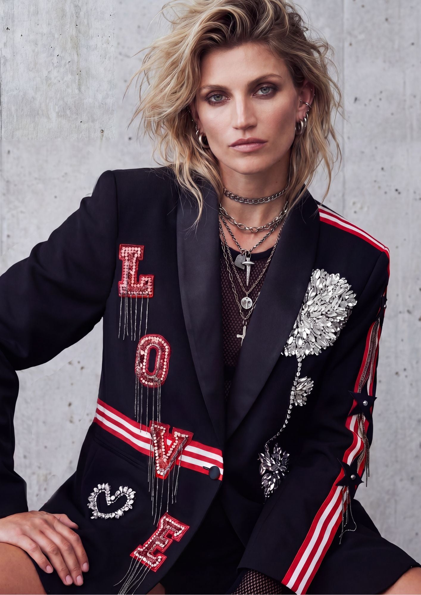 A model wearing a black blazer with 'LOVE' written in red and silver rhinestones, silver floral embellishments, and red and white striped details on the sleeves, posed against a textured concrete wall.