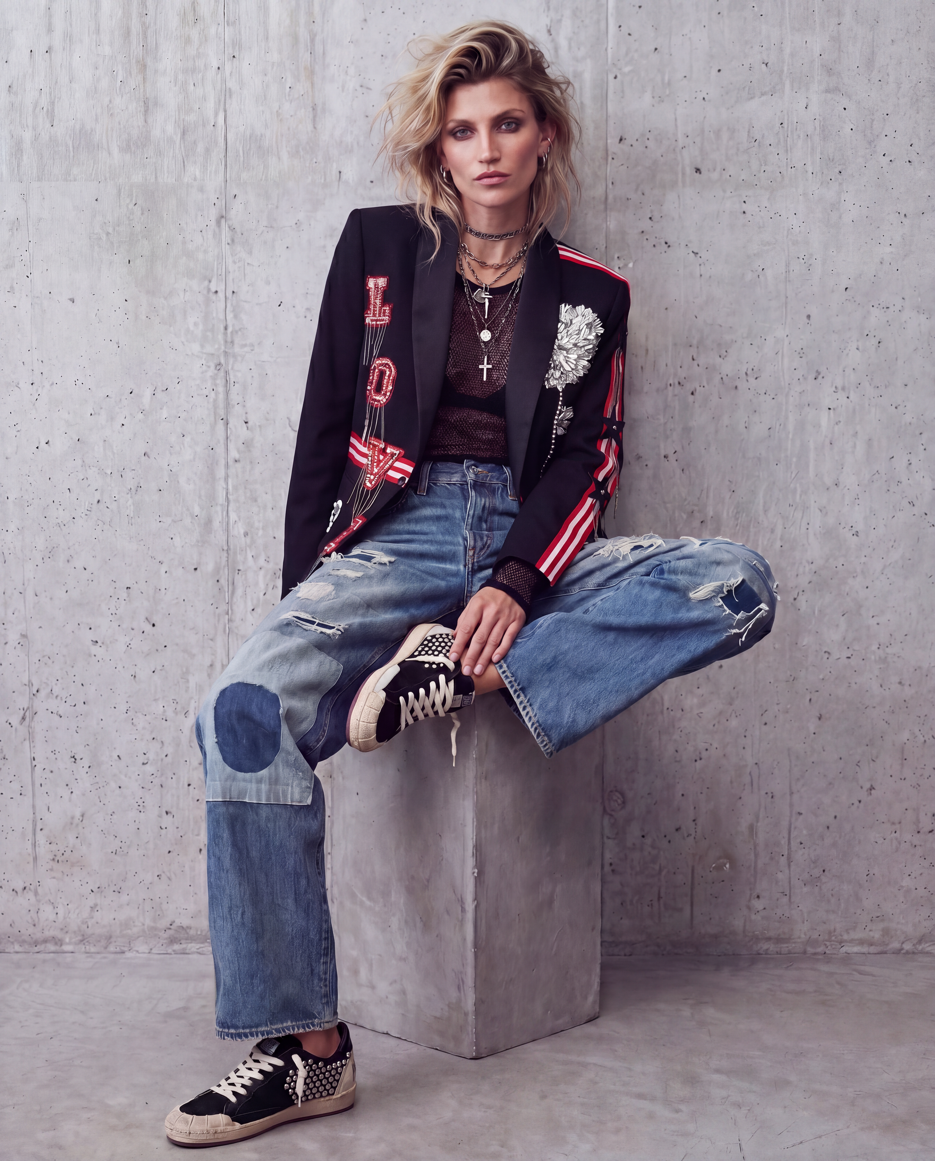 A model sits on a concrete block against a textured gray wall, wearing a black embroidered blazer with red and white accents, a dark brown top, distressed blue jeans with patchwork details, and black-and-white sneakers.