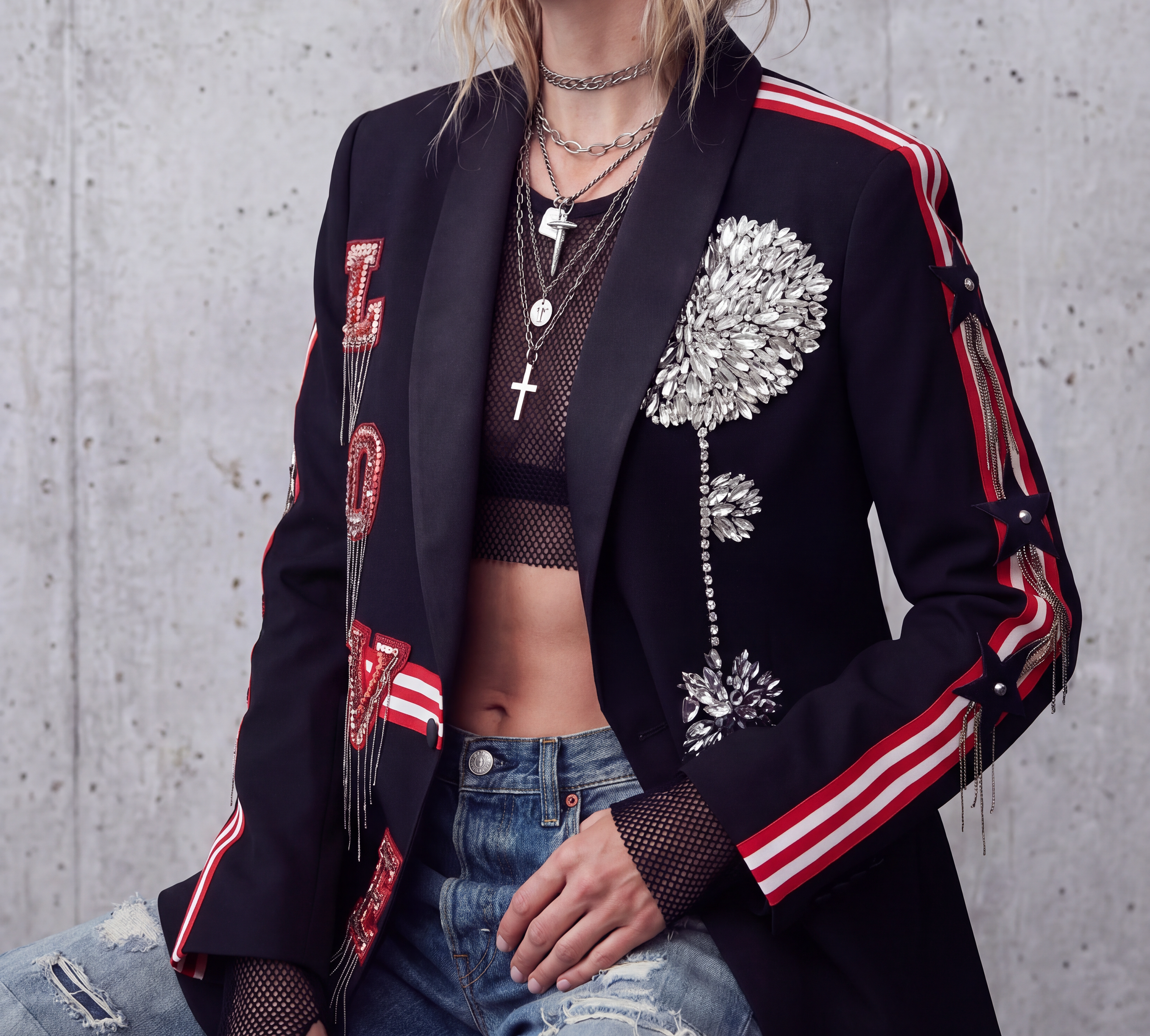 A person wearing a black tailored blazer with red and white striped sleeve details, silver floral embellishments, and red sequined lettering, paired with a black fishnet crop top and distressed blue jeans against a concrete wall.