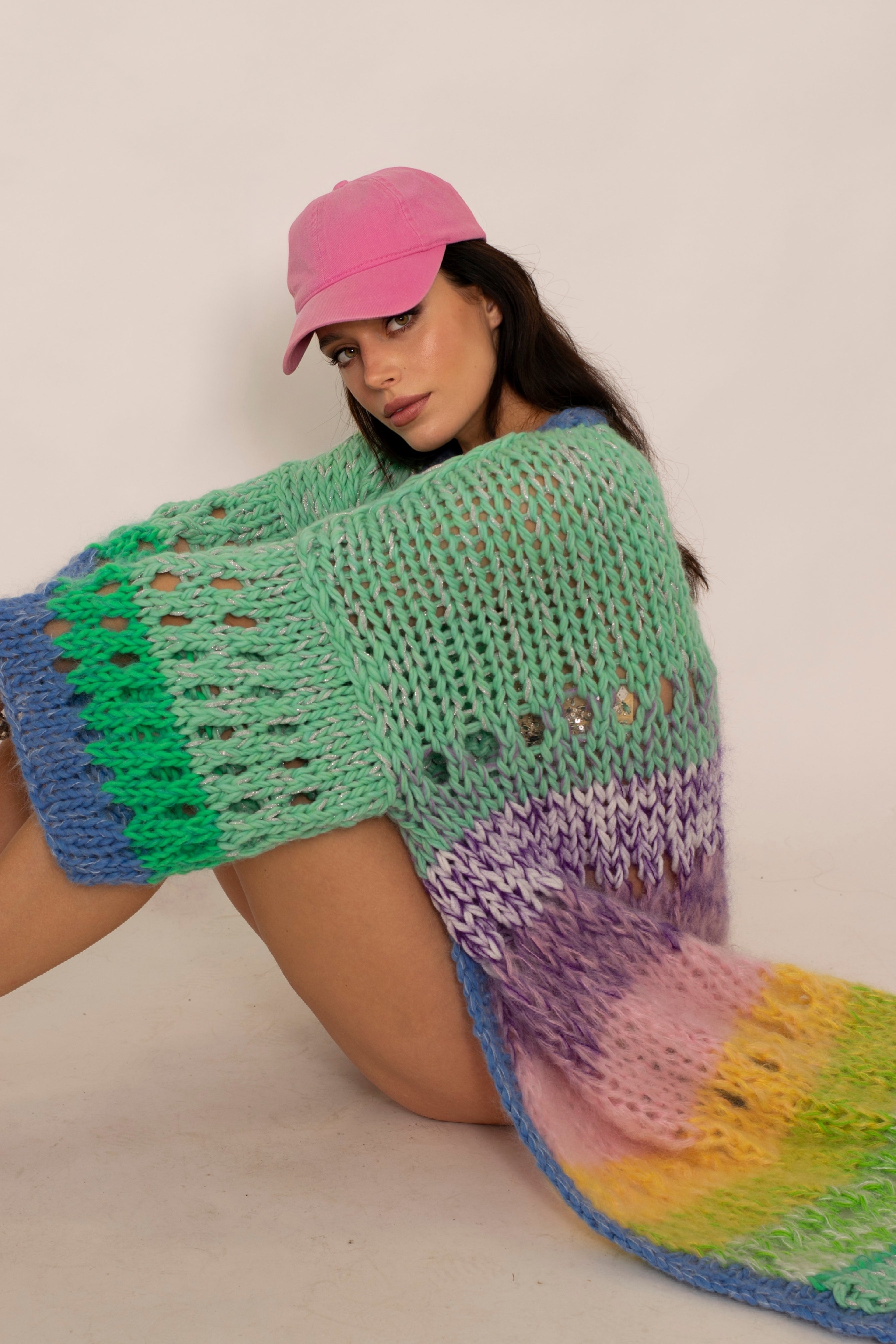 Girl with pink cap sitting on the floor wearing a pastel multicolor striped openwork long coat