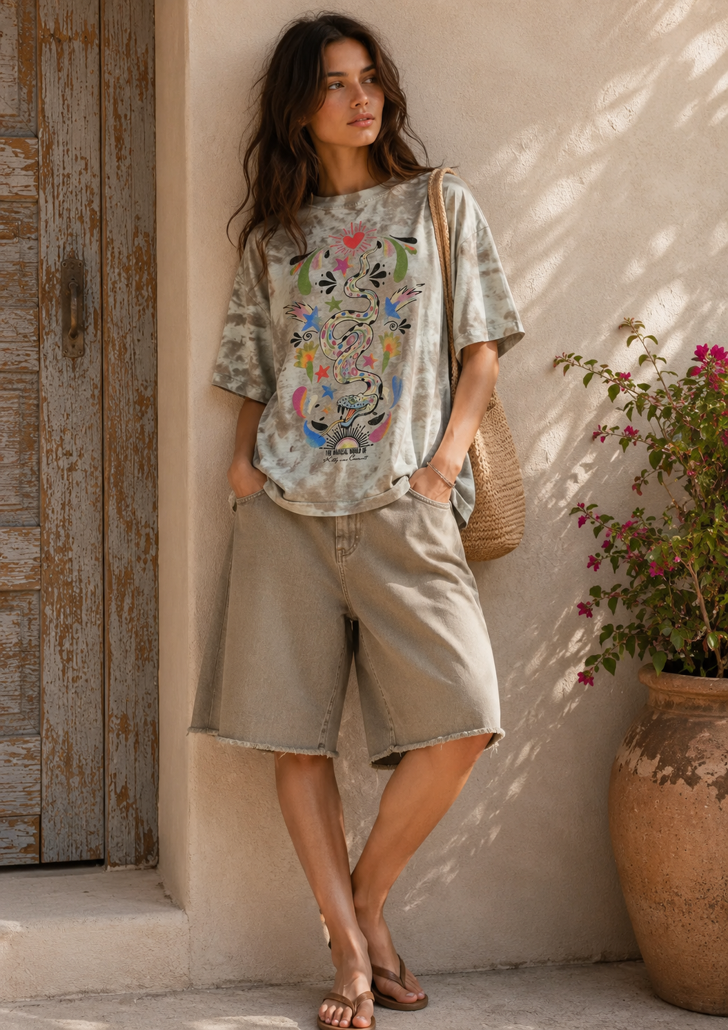 A woman stands against a sunlit stucco wall next to a rustic wooden door and a large potted plant, wearing a loose short-sleeve graphic T-shirt with a colorful floral and bird print, light beige shorts, and flat sandals. She is carrying a woven shoulder bag.
