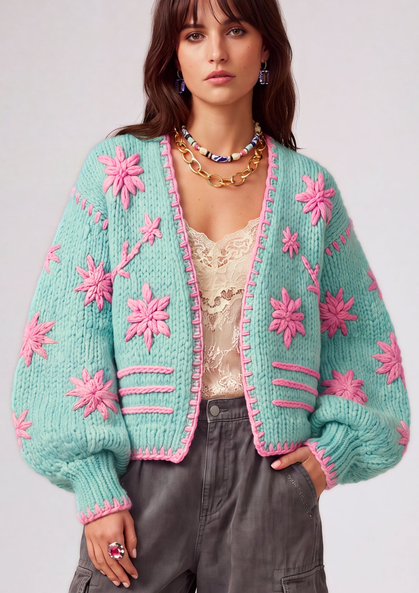 A woman wearing a mint green chunky knit cardigan with bright pink embroidered floral motifs and pink trim, styled over a cream lace top and dark gray cargo pants.