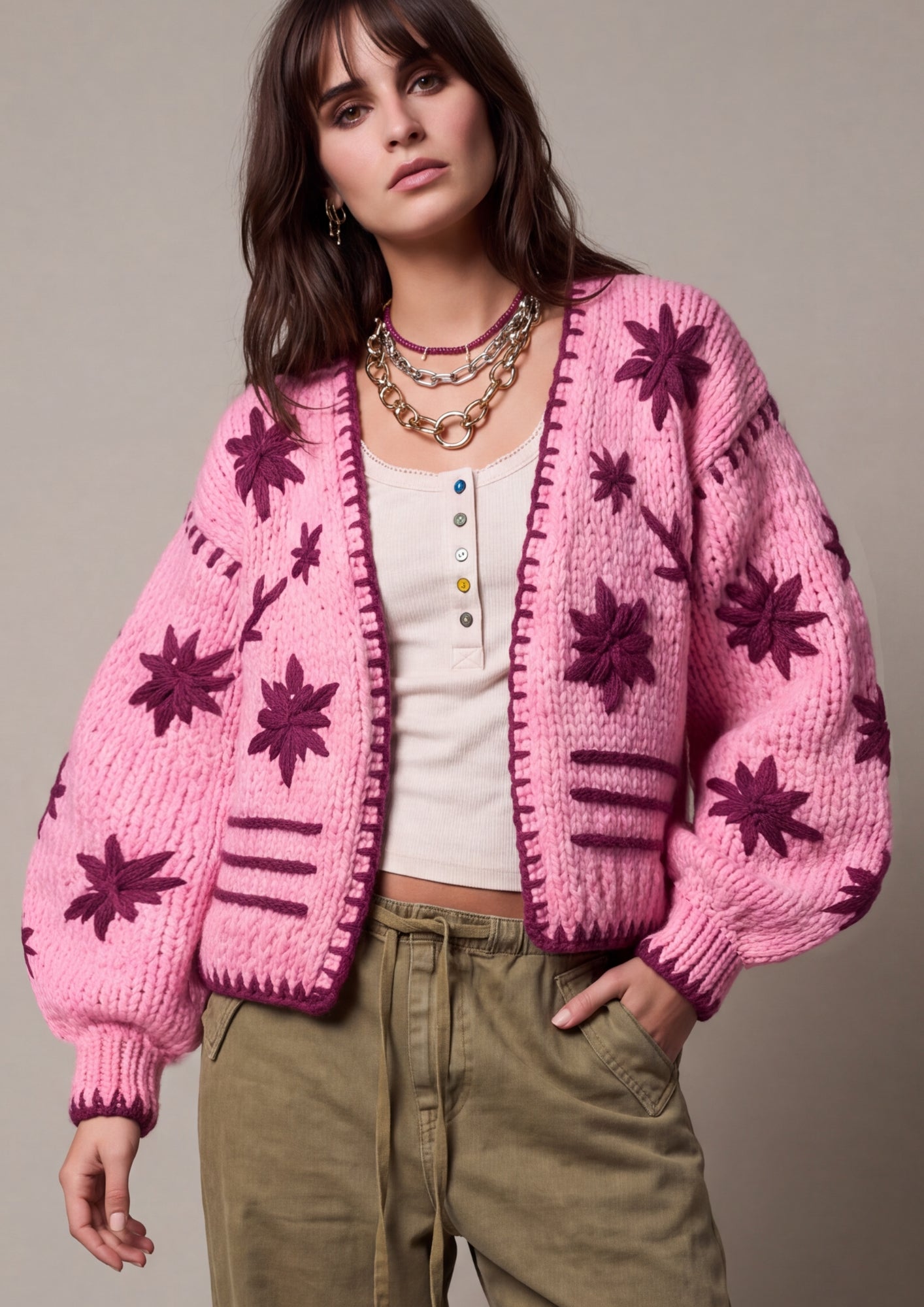 A woman models a pink chunky knit cardigan with dark purple starburst and stripe patterns, worn over a white top and olive green pants against a neutral studio background.