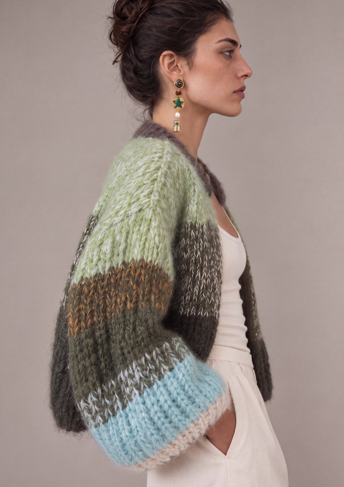 A woman in profile wearing a chunky knit cardigan with horizontal stripes in sage green, olive green, brown, light blue, and cream, paired with a white dress against a neutral gray background.