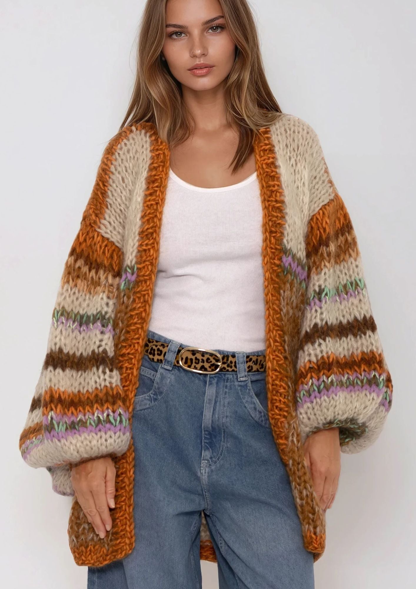 Woman wearing a patterned striped handknittedcardigan, in beige terra tones,white top, and blue jeans on a plain background