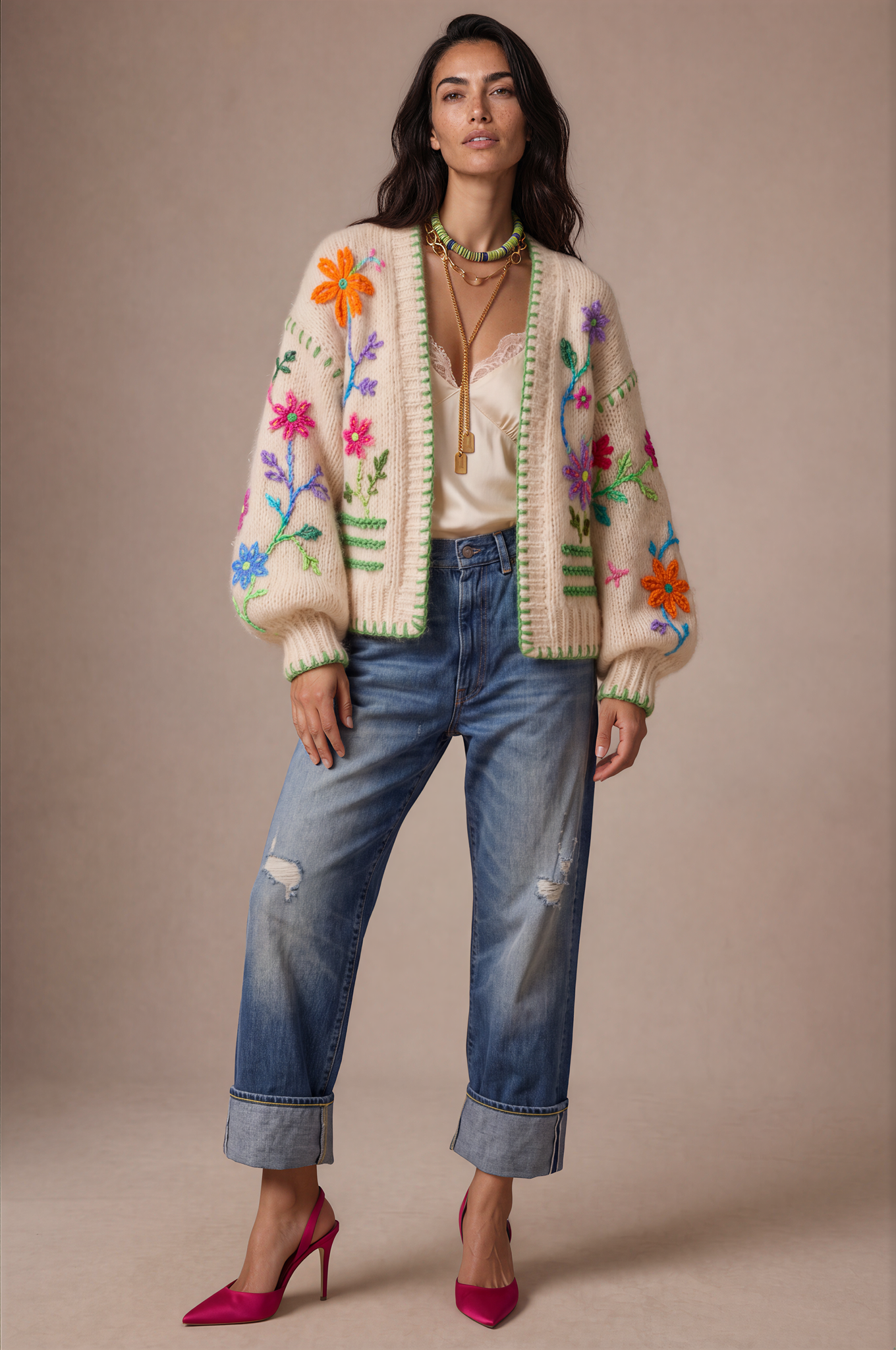 A model stands against a solid beige studio background wearing a cream-colored knit cardigan with colorful floral embroidery, blue distressed jeans, and pink heels.