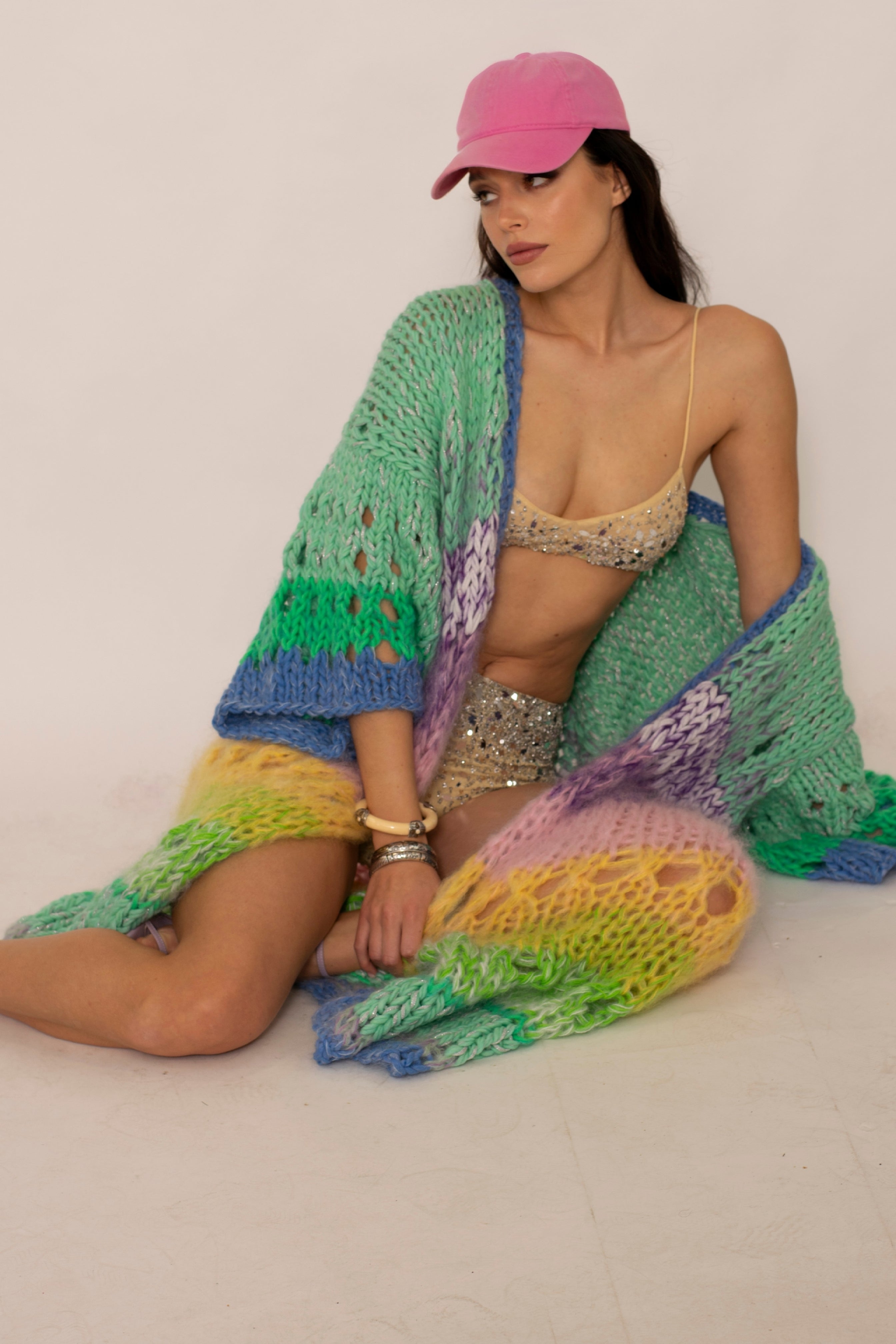 girl sitting on the ground wearing a sandcolor bikini with sequins and a pastel colorful handknitted long cardigan