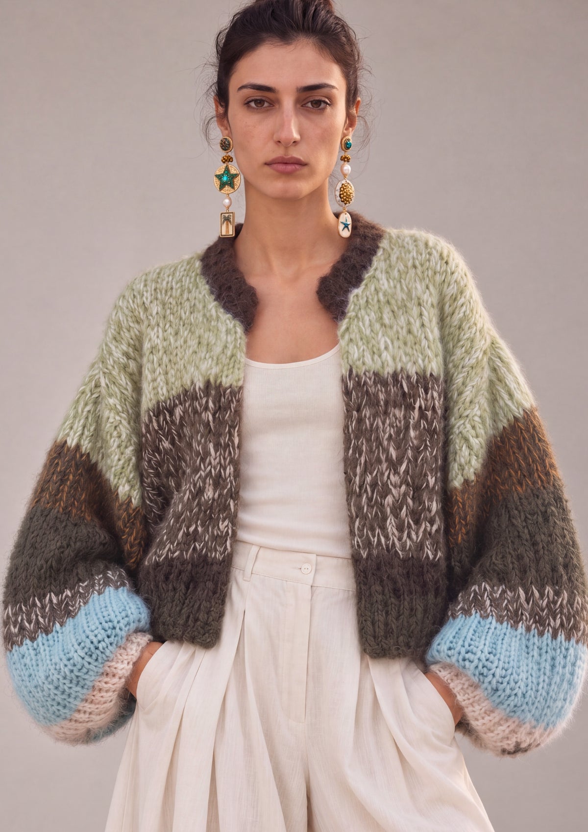 A woman models a chunky knit cardigan with color-blocked stripes in sage green, brown, beige, and light blue, styled over a white tank top and cream wide-leg trousers against a neutral studio background.