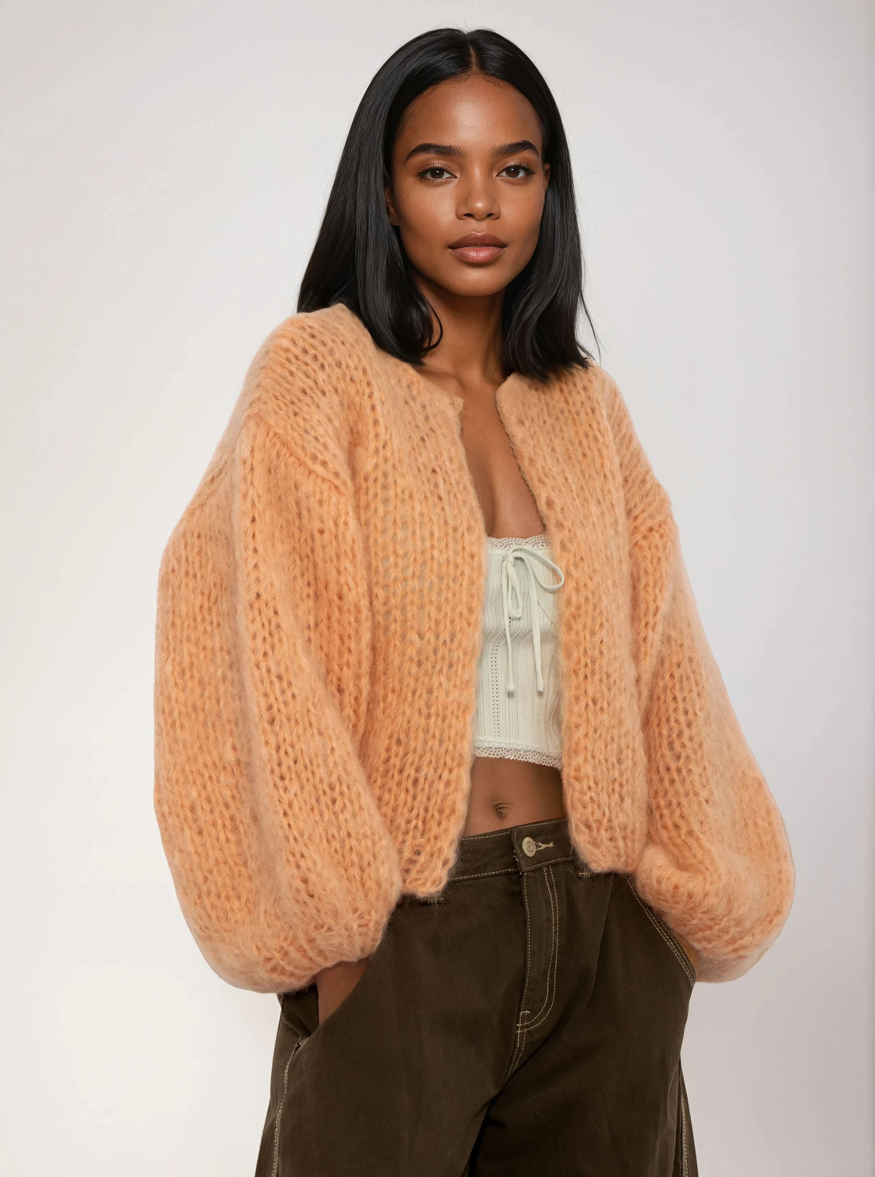 Woman wearing a light orange cardigan over a white top and brown pants on a plain background