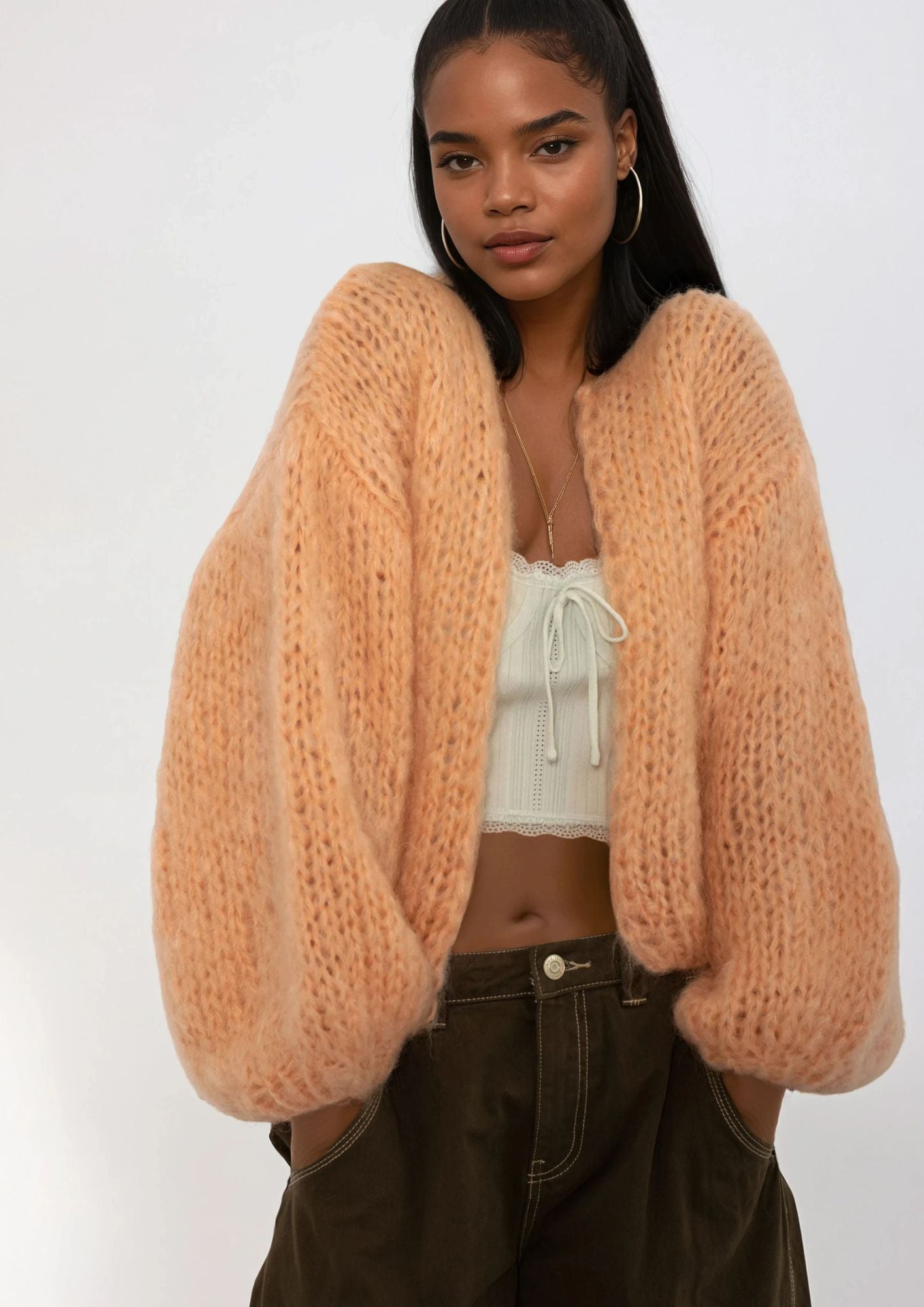 Woman wearing a mohair orange chunky knit cardigan over a white top and dark pants on a white background