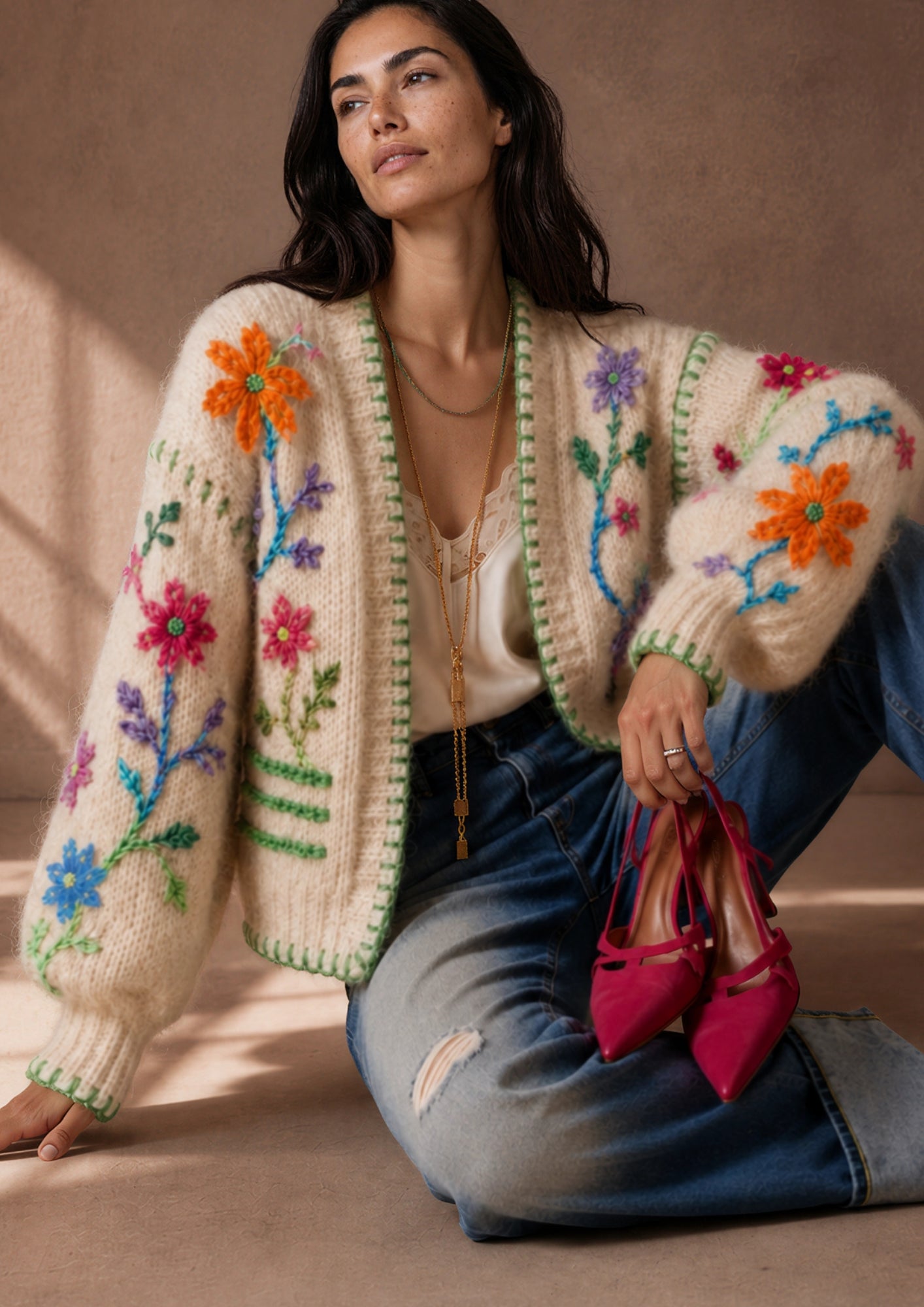 A woman sits on a beige floor wearing a cream knit cardigan with colorful floral embroidery, blue jeans, and holding a pair of bright pink pointed-toe heels.