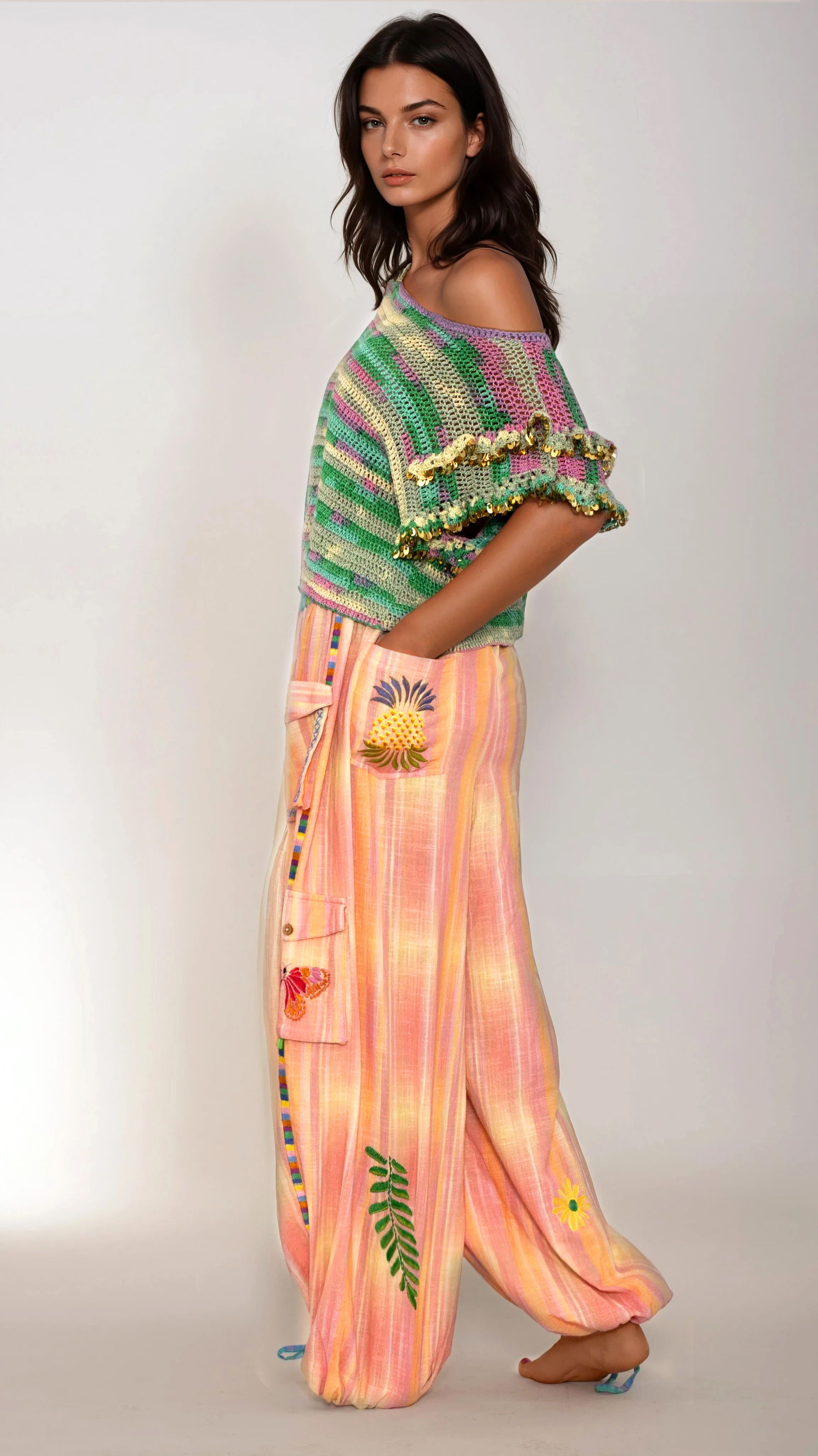 Woman wearing a colorful crochet top with sequins and tie-dye pants with pineapple designs  embroidery on a plain background