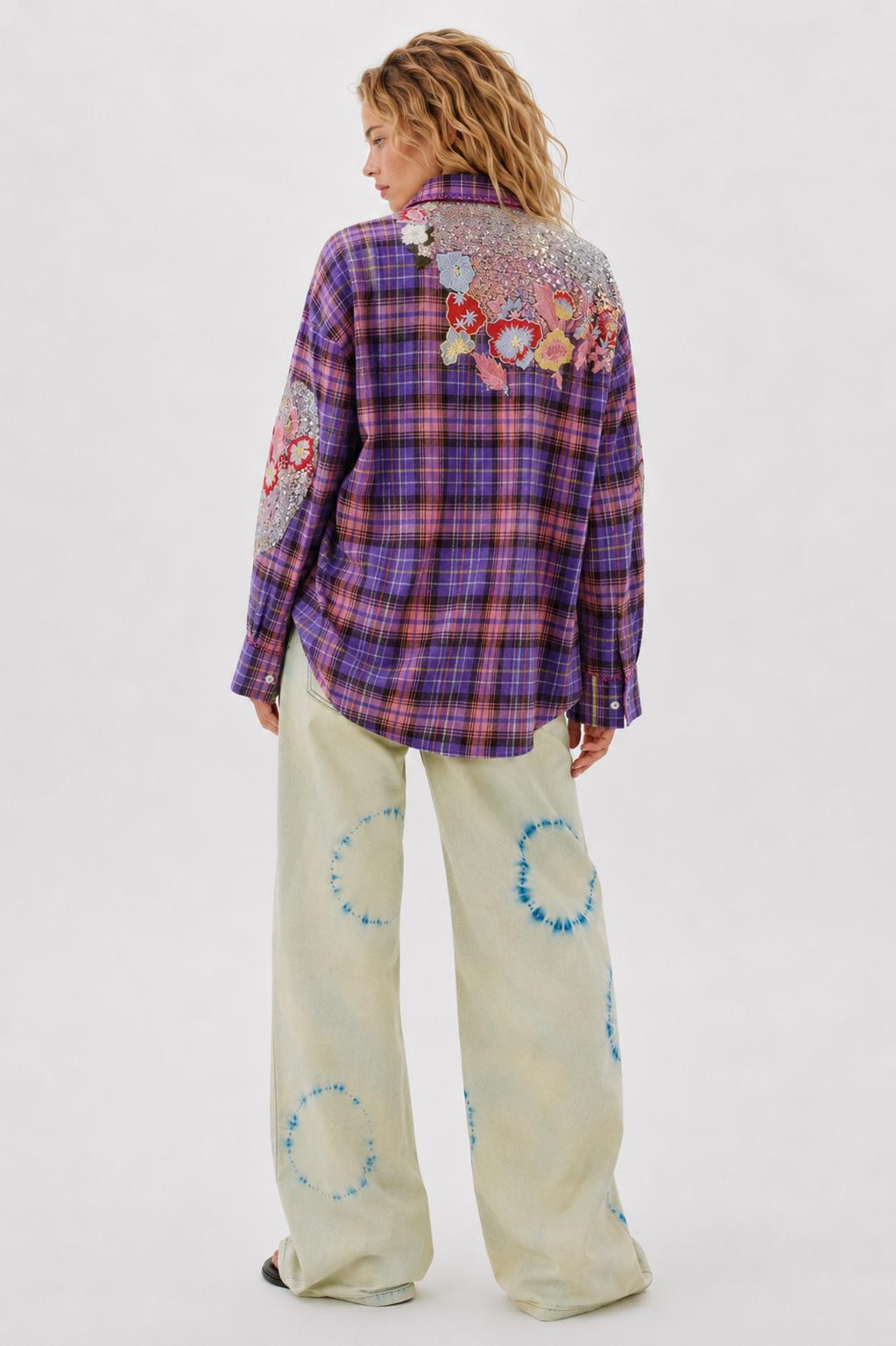 Person wearing a plaid shirt with floral embroidery and patterned pants on a white background
