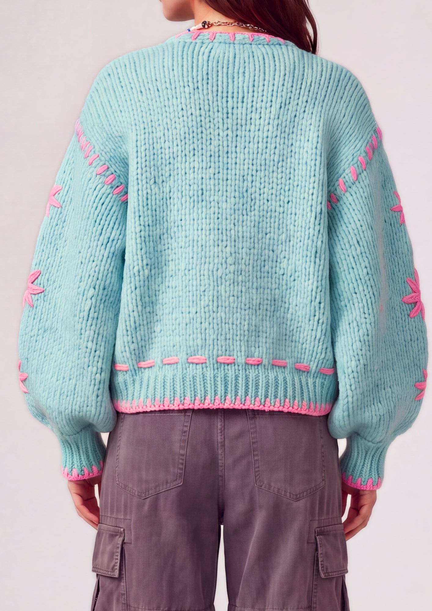 A person shown from the back wearing a chunky knit sweater in light aqua with bright pink contrast stitching and pom-pom sleeves, paired with gray cargo pants against a plain light background.