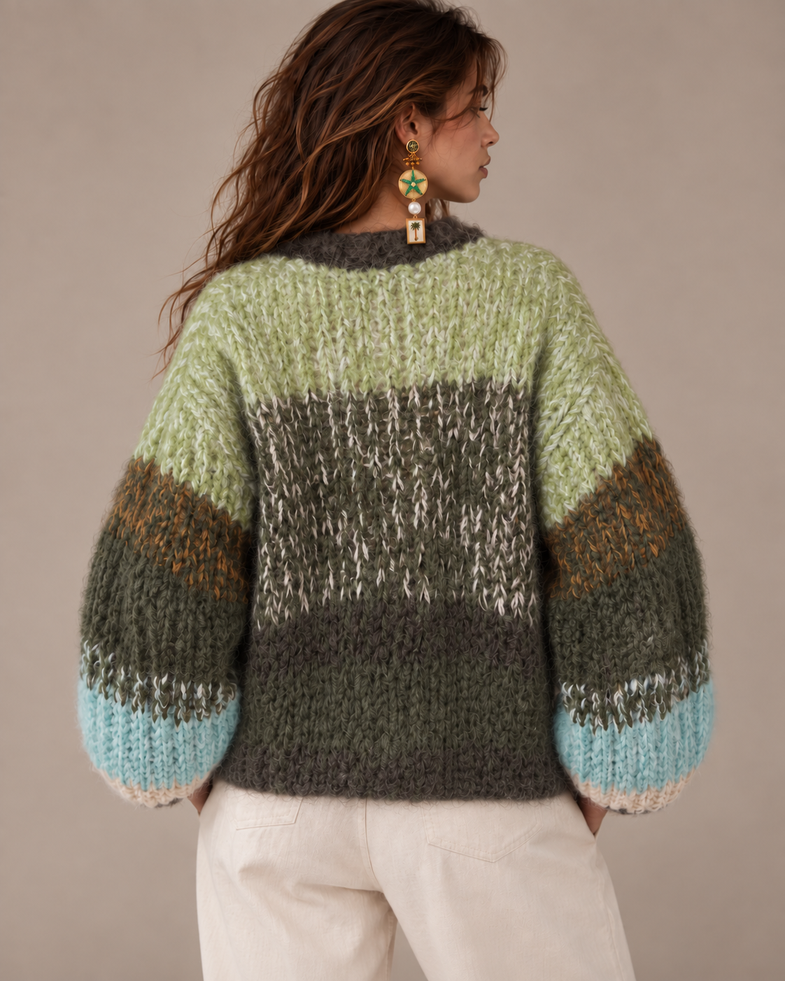 A woman shown from the back wearing a chunky knit sweater with horizontal color-blocked stripes in sage green, olive green, brown, and light blue, paired with cream pants against a neutral beige background.