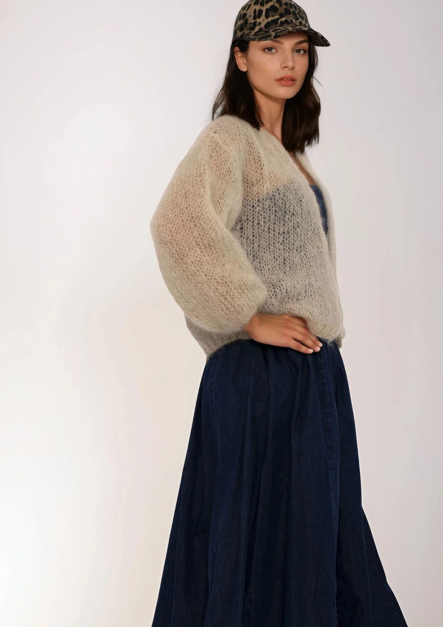 Woman wearing a beige handknit sweater and navy skirt with a leopard print cap on a white background