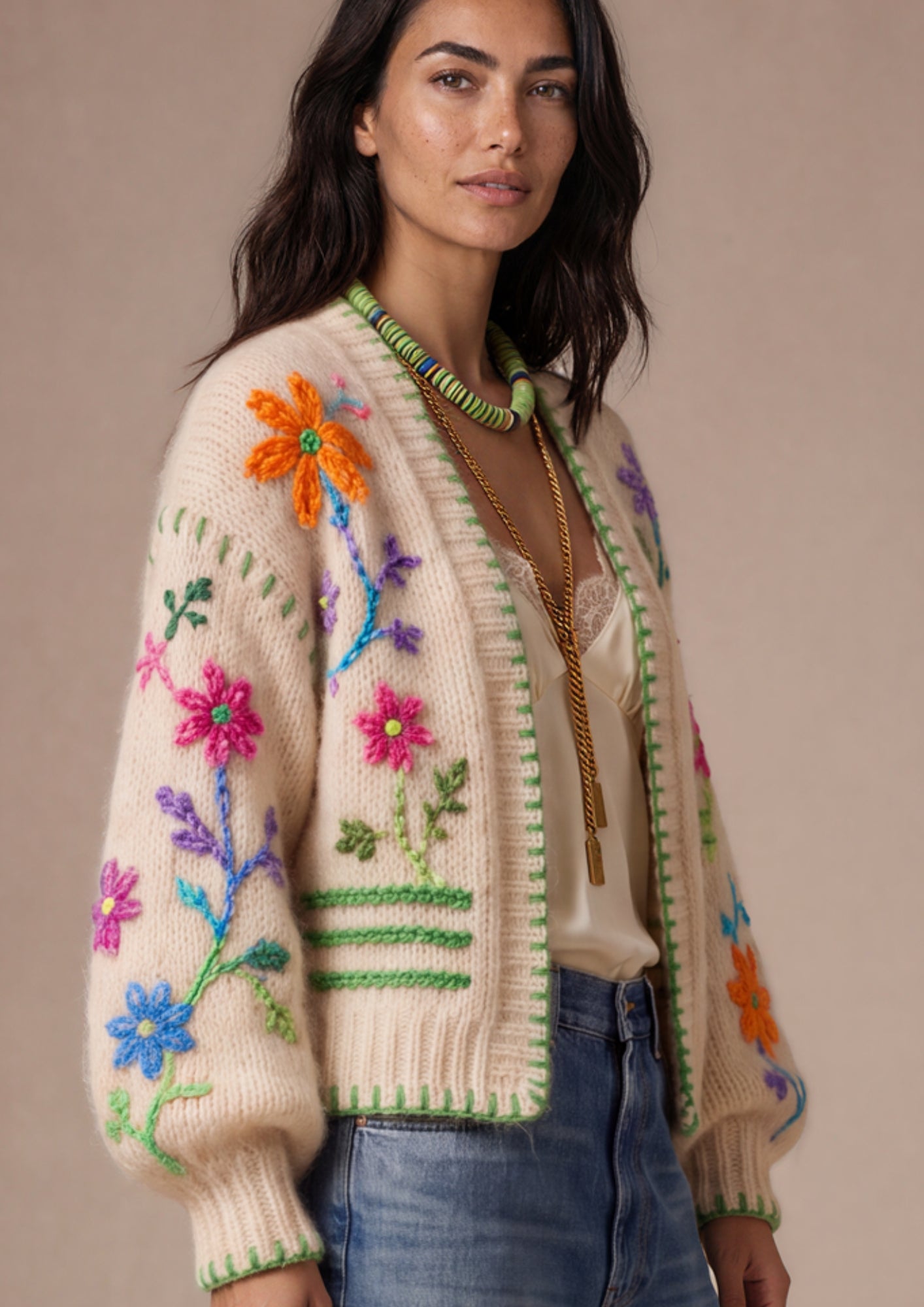 A woman wearing a cream-colored knit cardigan with colorful hand-knitted floral patterns and green trim, paired with blue jeans and gold jewelry, set against a solid beige background.