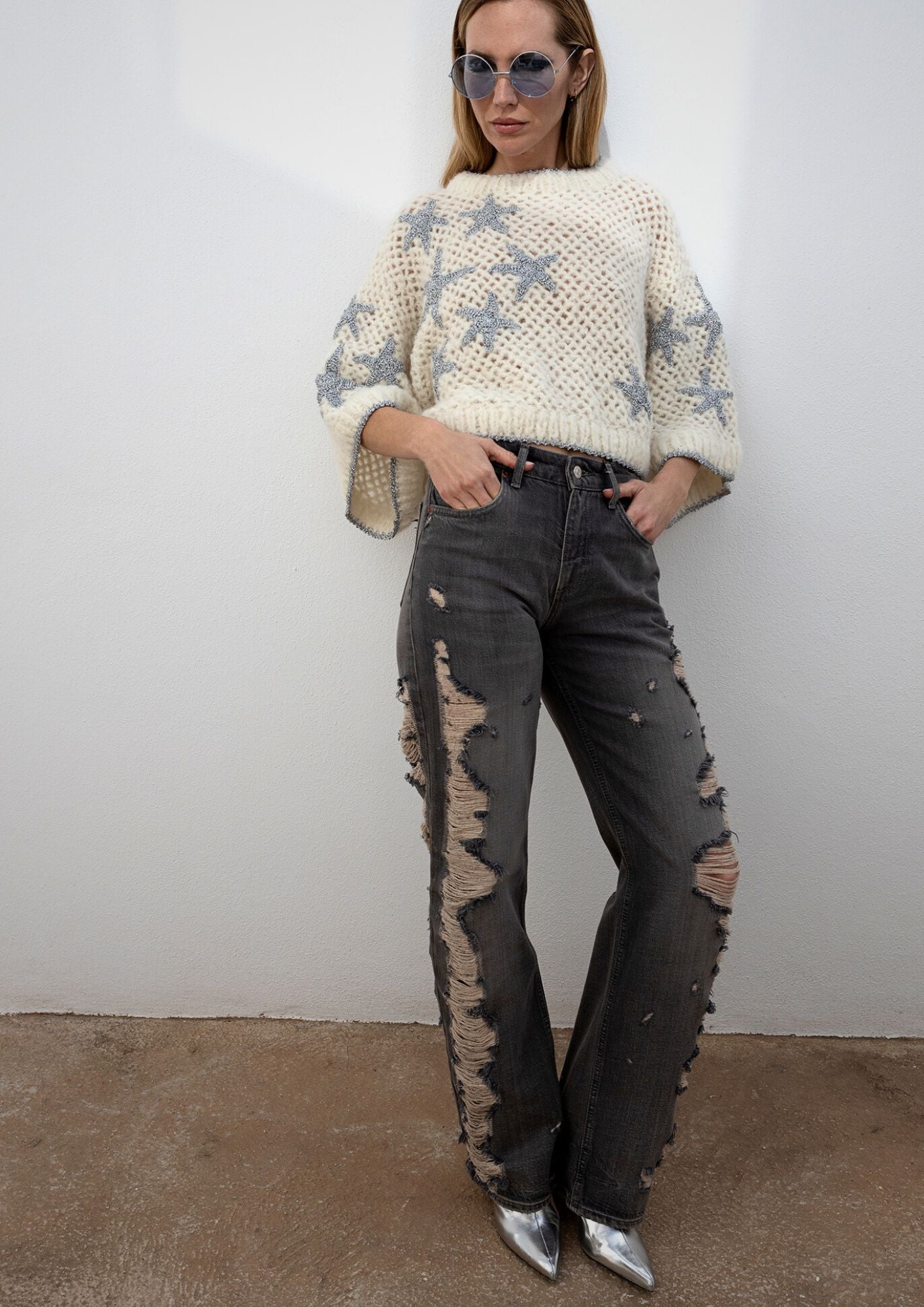 Girl leaning agianst a wall wearing a crocheted creme top, with vintage jeans