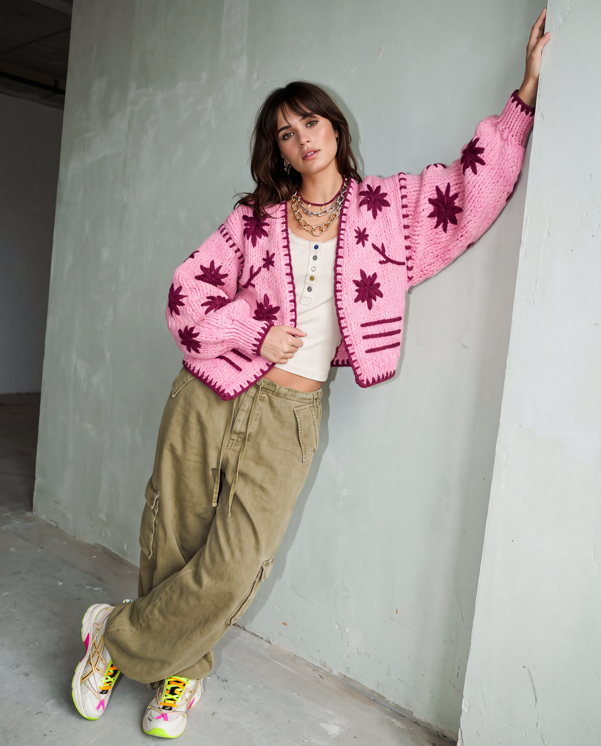 A woman posing in a pink and purple patterned knit cardigan, olive cargo pants, and colorful sneakers, leaning against a concrete wall.