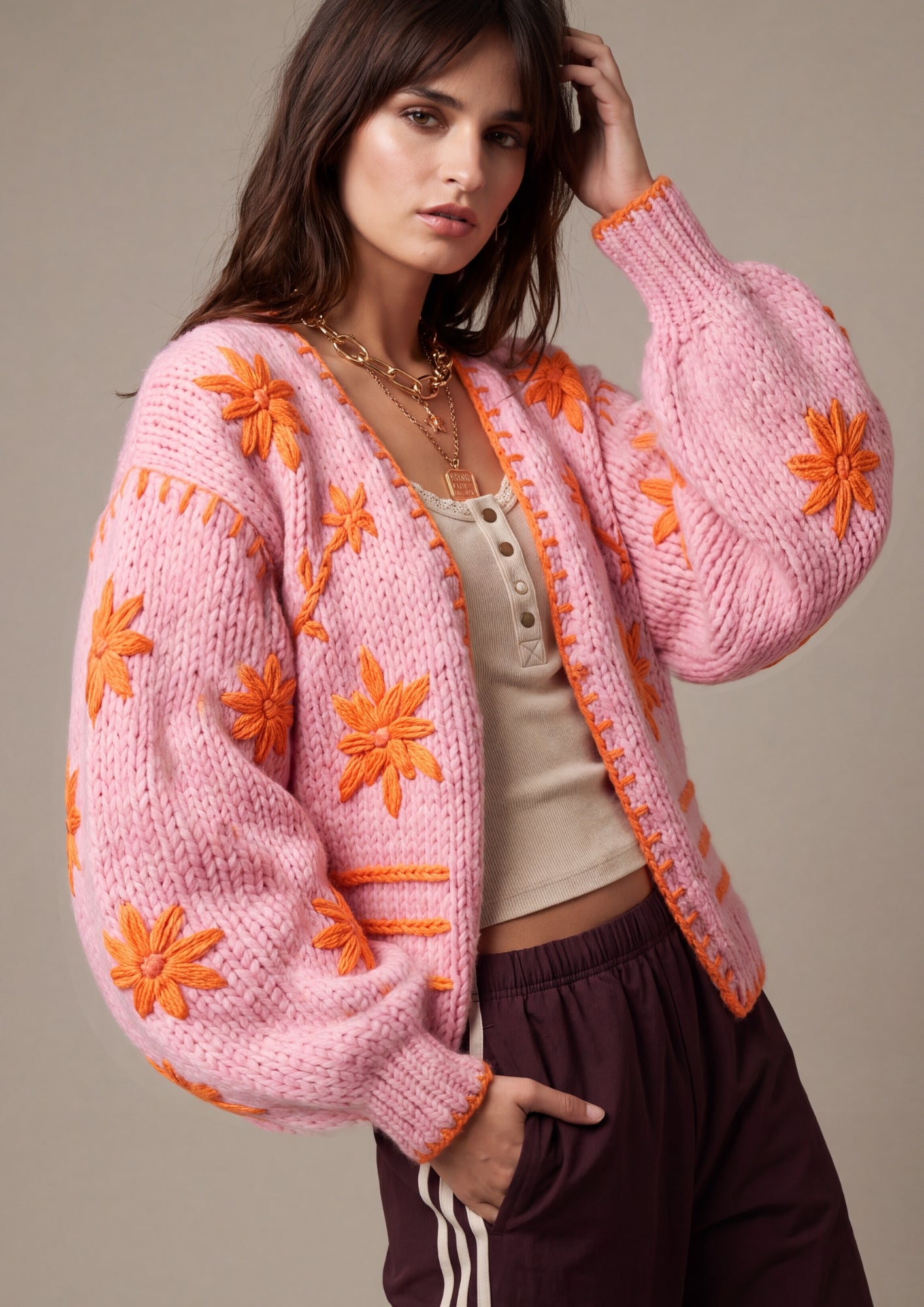 A woman models a chunky pink knit cardigan with bright orange embroidered floral motifs and orange trim, worn over a beige top and dark brown sweatpants against a solid beige studio background.
