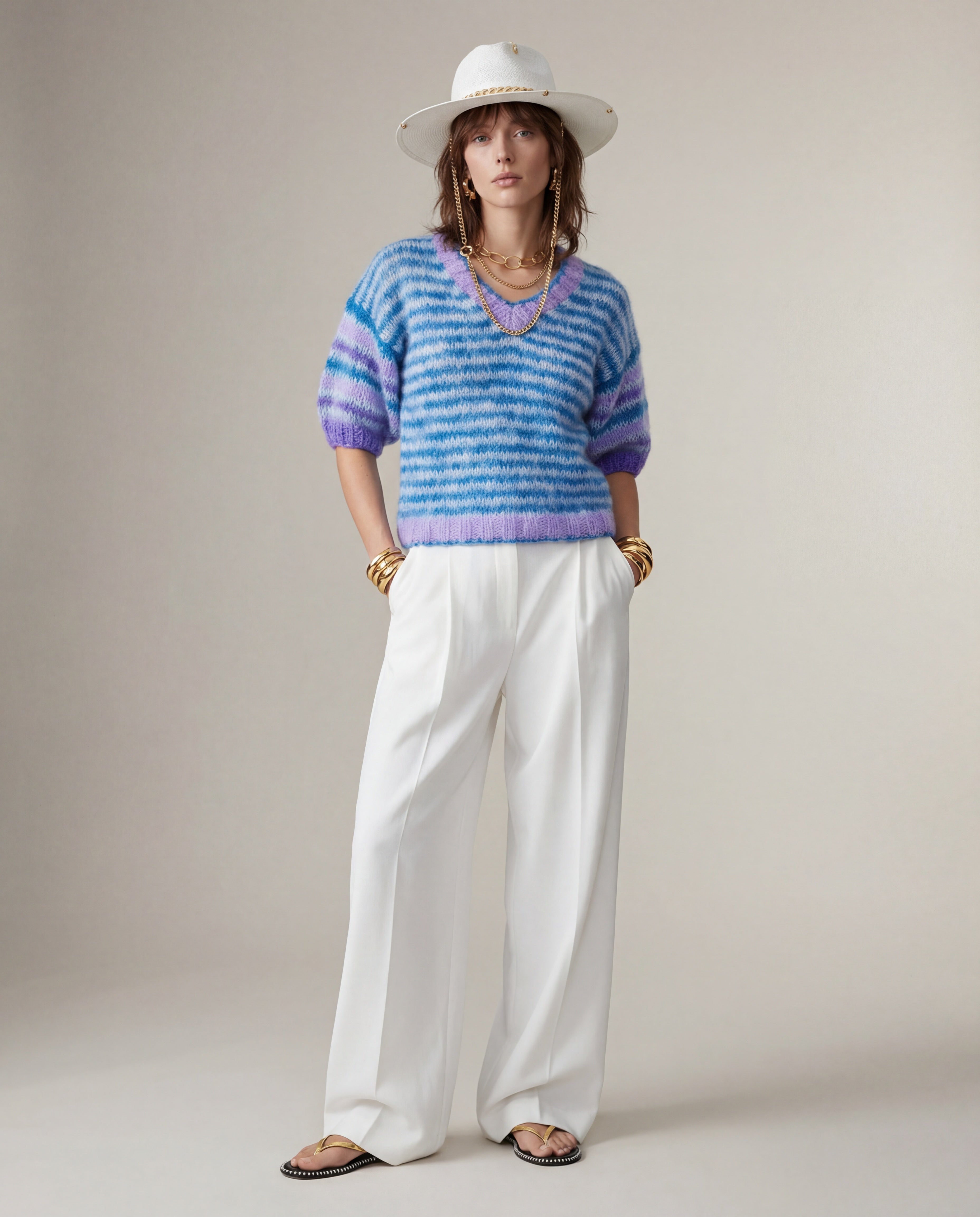 Woman wearing a blue and purple striped sweater with white pants and a white hat on a plain background